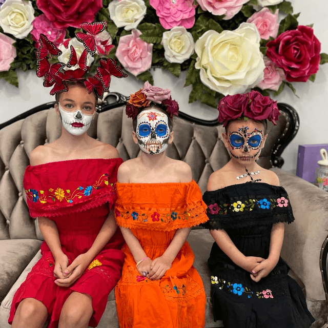 Three girls in colorful traditional dresses and Day of the Dead face paint sitting on a couch with large paper flowers backdrop