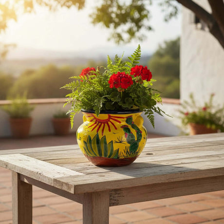 The Talavera Hub yellow ceramic planter with hand-painted desert cactus and sun design. Holds red flowers and green ferns.