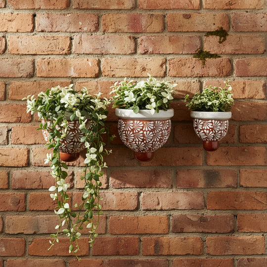 The Talavera Hub set of three brown and white hand-painted ceramic wall planters with floral patterns on a brick wall.