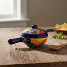 The Talavera Hub hand-painted ceramic salsa bowl with lid, featuring multicolor floral and geometric patterns on a yellow base with a blue rim and handles.