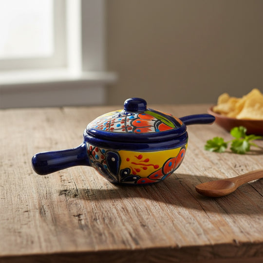 The Talavera Hub hand-painted ceramic salsa bowl with lid, featuring multicolor floral and geometric patterns on a yellow base with a blue rim and handles.