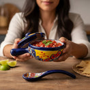 The Talavera Hub hand-painted ceramic salsa bowl with lid and spoon, featuring multicolor floral and geometric patterns on a yellow and blue base.