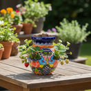 The Talavera Hub hand-painted Mexican ceramic strawberry planter with vibrant floral patterns on a wooden garden table.