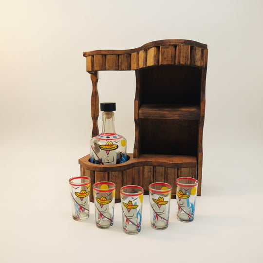 The Talavera Hub Mexican Tequila Decanter Set with 6 shot glasses on a rustic wooden stand. Hand-painted figures in sombreros adorn the glassware.