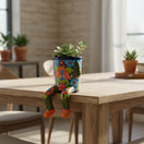 The Talavera Hub hand-painted ceramic planter with arms and legs, featuring colorful floral designs and a blue base, sits on a table edge.
