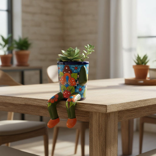 The Talavera Hub hand-painted ceramic planter with arms and legs, featuring colorful floral designs and a blue base, sits on a table edge.