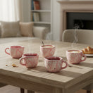 The Talavera Hub set of four hand-painted pink ceramic mugs with white floral patterns on a wooden dining table.