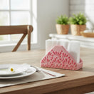 The Talavera Hub pink ceramic napkin holder with embossed floral design, holding white napkins on a wooden table.