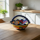The Talavera Hub hand-painted ceramic fruit basket with blue handles, yellow rim, and vibrant floral patterns in red, orange, and blue.