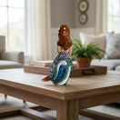 The Talavera Hub hand-painted ceramic mermaid figurine with floral tail, holding a yellow fish, resting on blue and green waves.