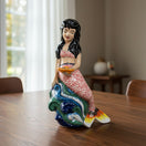 The Talavera Hub hand-painted ceramic mermaid figurine with pink floral tail, coral top, black hair, holding a yellow fish on blue and green waves.